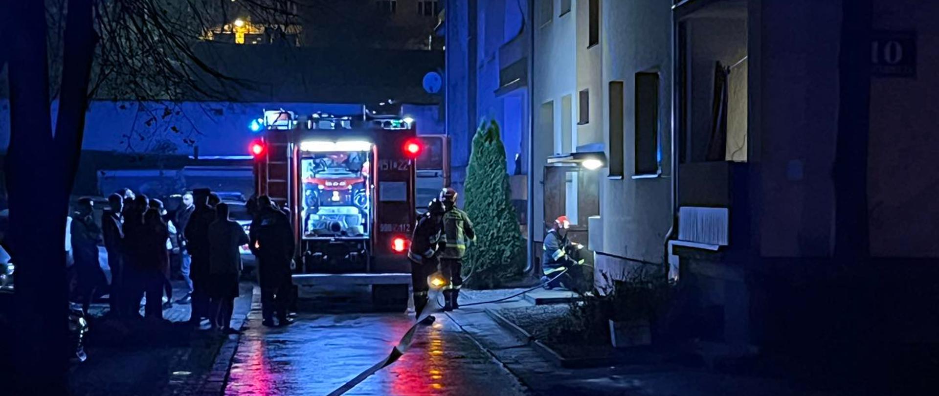 Na zdjęciu w porze nocnej widać na pierwszym planie samochód strażacki zaparkowany przed blokiem, w którym doszło do pożaru. Samochód zaparkowany tyłem do zdjęcia. Przy samochodzie widać strażaków, którzy prowadzą akcją gaśniczą oraz tłum gapiów.