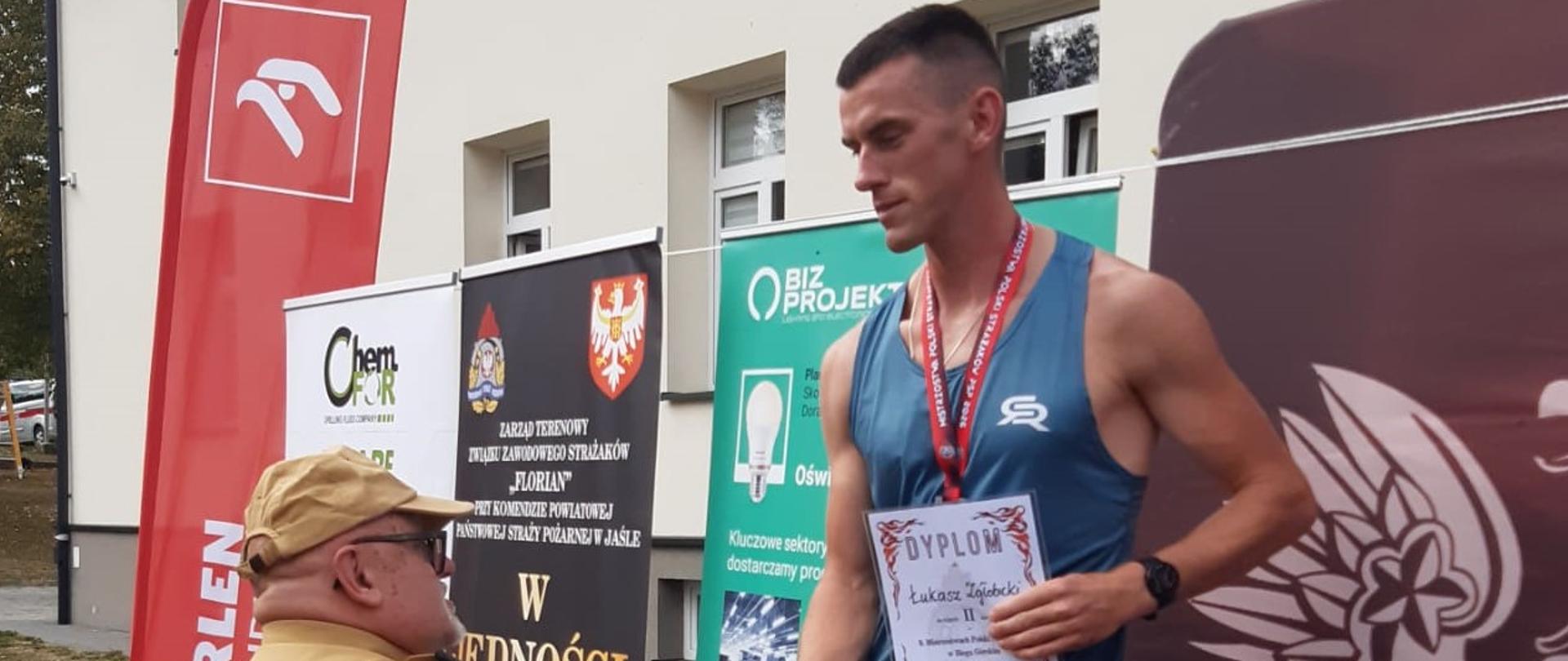Moment wręczania nagród dla uczestników biegu. Zawodnik, który zajął drugie miejsce przyjmuje gratulacje od funkcjonariusza PSP, który ubrany jest w ubranie piaskowe. Zawodnik trzyma w ręku dyplom, na szyi widać czerwoną smycz, na której wisi medal. Po prawej stronie zdjęcia widać część banneru PSP, po lewej stronie widać bannery reklamowe sponsorów zawodów. 
