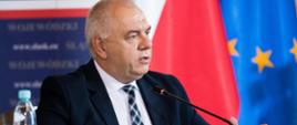 Wicepremier Jacek Sasin przemawia siedząc za stołem. W tle flaga Polski i Unii Europejskiej.