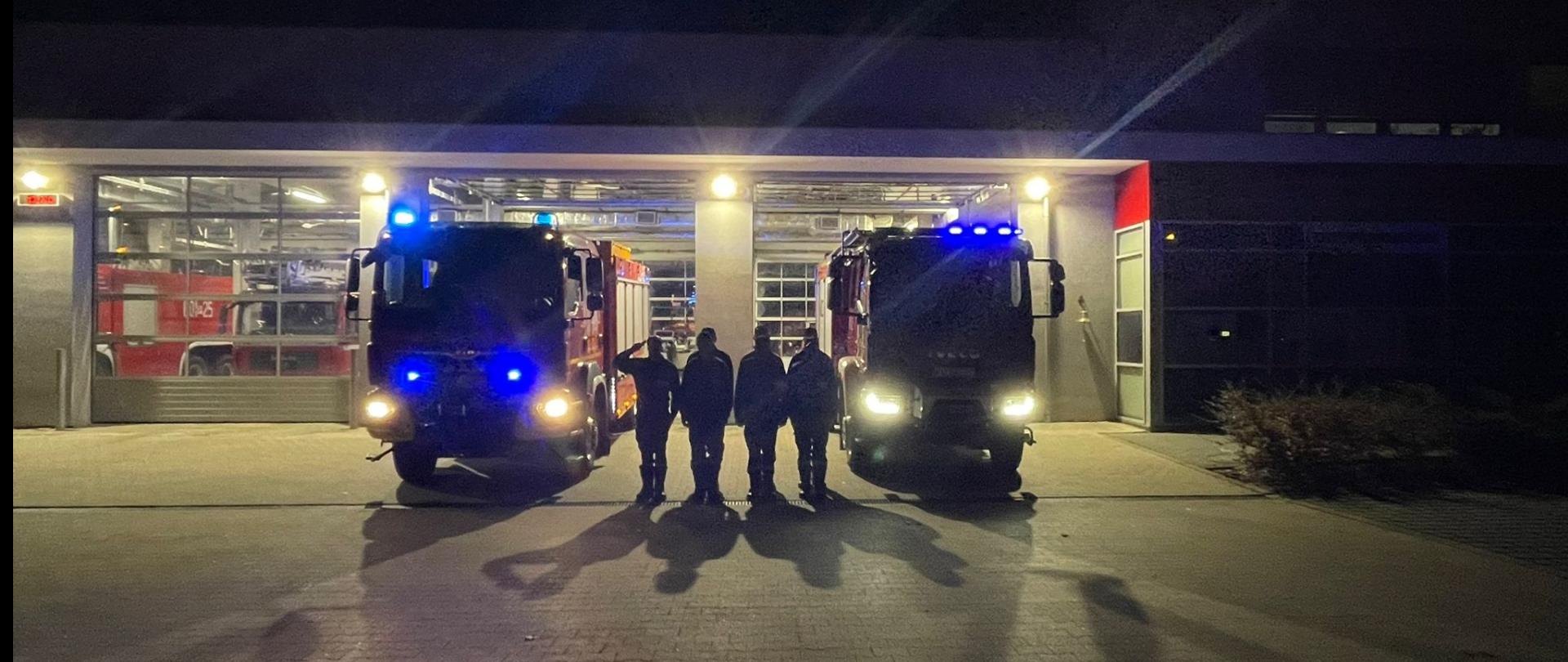 Na zdjęciu stoją strażacy przed budynkiem KP PSP Śrem oraz pojazdami ratowniczo gaśniczymi z włączonymi sygnałami dźwiękowymi i świetlnymi oddając hołd zmarłemu strażakowi z JRG 2 Gdańsk