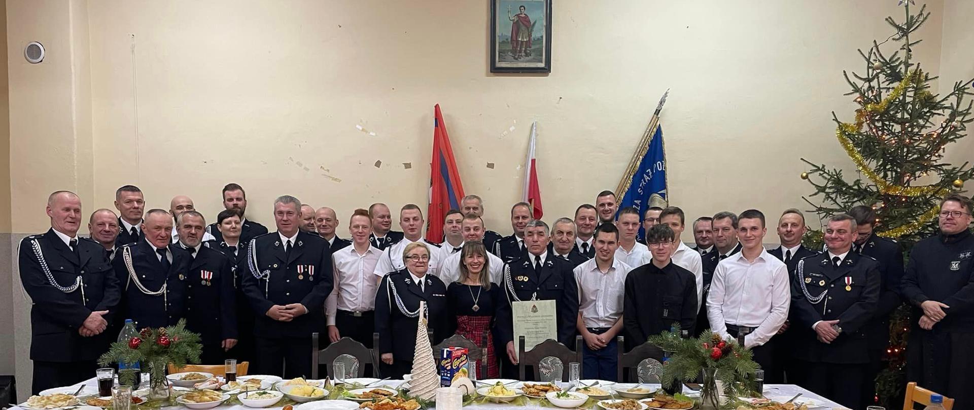 Na zdjęciu widać posła na sejm w umundurowaniu OSP oraz druhów i duhny OSP - wszyscy w mundurach wyjściowych. Prezes OSP trzyma w ręku decyzję o włączeniu do KSRG. W tle flagi Polski oraz związku OSP RP.. Przed strażakami stół w typowym wigilijnym nakryciu. 