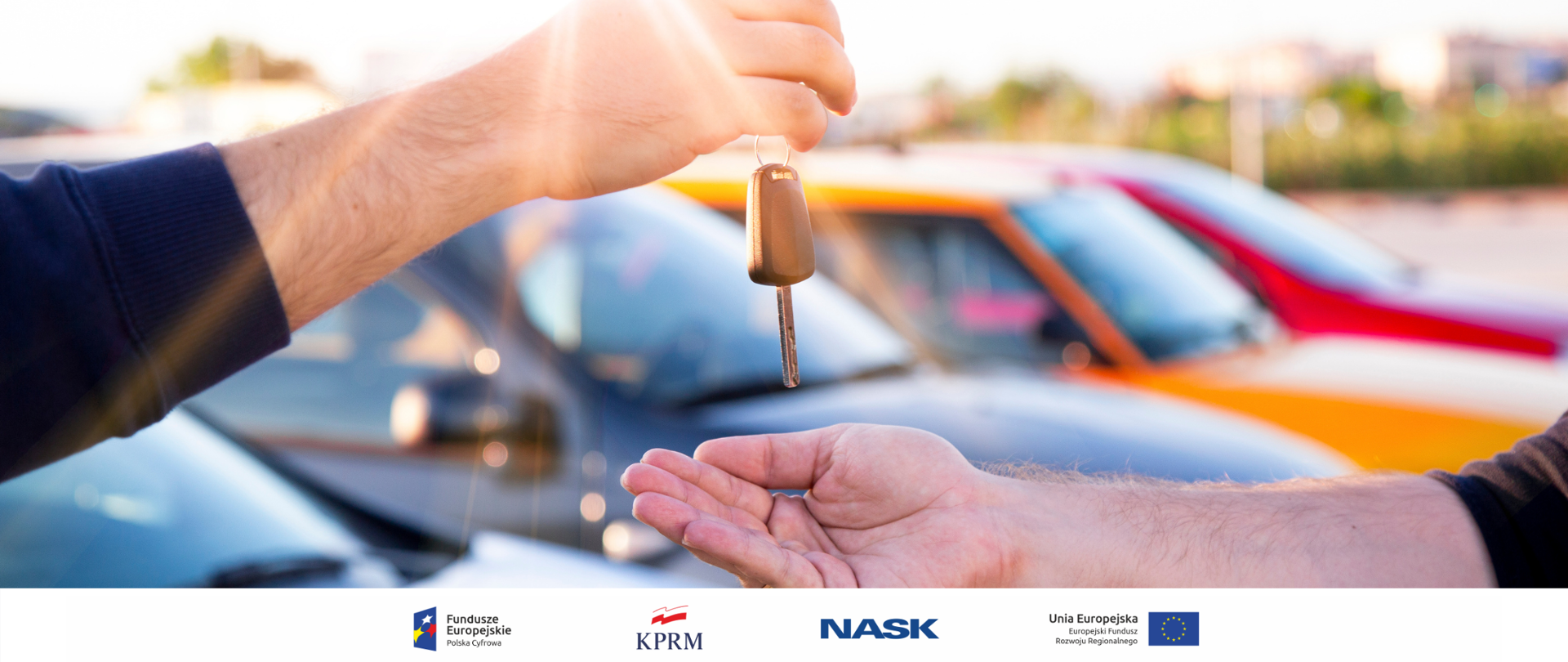 Zdjęcie męskich rąk. Jeden przekazuje drugiemu kluczyki do samochodu. W tle widoczne kolorowe auta.