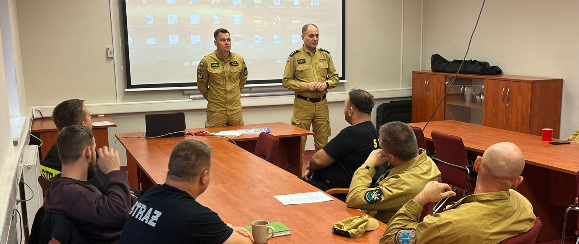 Na zdjęciu widać grupę osób strażaków siedzących w sali szkoleń w jednostce ratowniczej-gasniczej. Przy długim, drewnianym stole siedzi kilkunastu uczestników szkolenia kierowców konserwatorów OSP, ubranych w mundury w kolorze żółtym oraz czarnym z napisem „STRAŻ”. Dwóch mężczyzn – komendant powiatowy starszy kapitan Mirosław Śledź oraz kierownik kursu, kapitan Konrad Okleciński w żółtych mundurach stoi z przodu sali, prowadząc szkolenie. Za nimi znajduje się ekran projekcyjny z wyświetlonym pulpitem komputera..