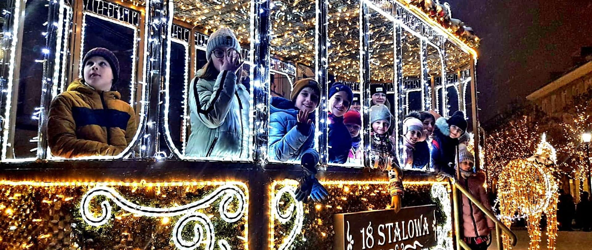 Uczniowie szkoły stoją w świętecznie oświetlonej dekoracji ulicznej w formie tramwaju z napisem 18 STALOWA na Krakowskim Przedmieściu w Warszawie