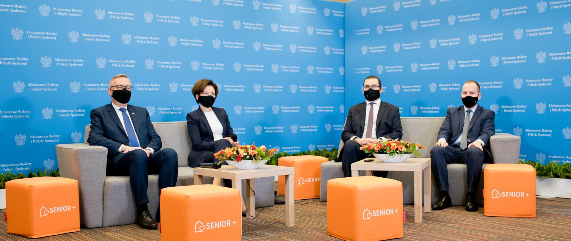 Uczestnicy konferencji (cztery osoby, w tym minister Adam Andruszkiewicz) siedzą w maseczkach na szarych kanapach. Przed nimi stoliki i pomarańczowe pufy z napisem Senior+.