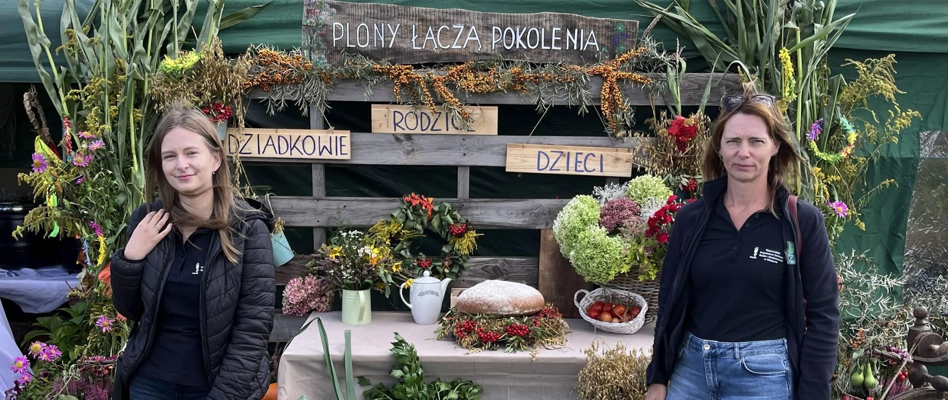 2 kobiety stojące na trawie, na tle wystawy z napisem plony łączą pokolenia dziadkowie rodzice dzieci, widoczny w tle stół a na nim bochen chleba, dzbanek, kubek, przed stołem w skrzynkach warzywa