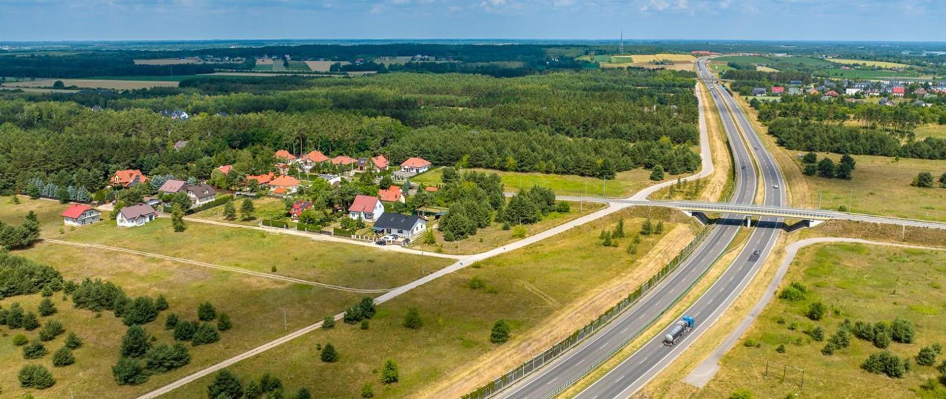 Widok z lotu ptaka na dwujezdniową drogę ekspresową przebiegającą przez tereny zielone, z wiaduktem drogowym oraz pobliską zabudową jednorodzinną i lasami.