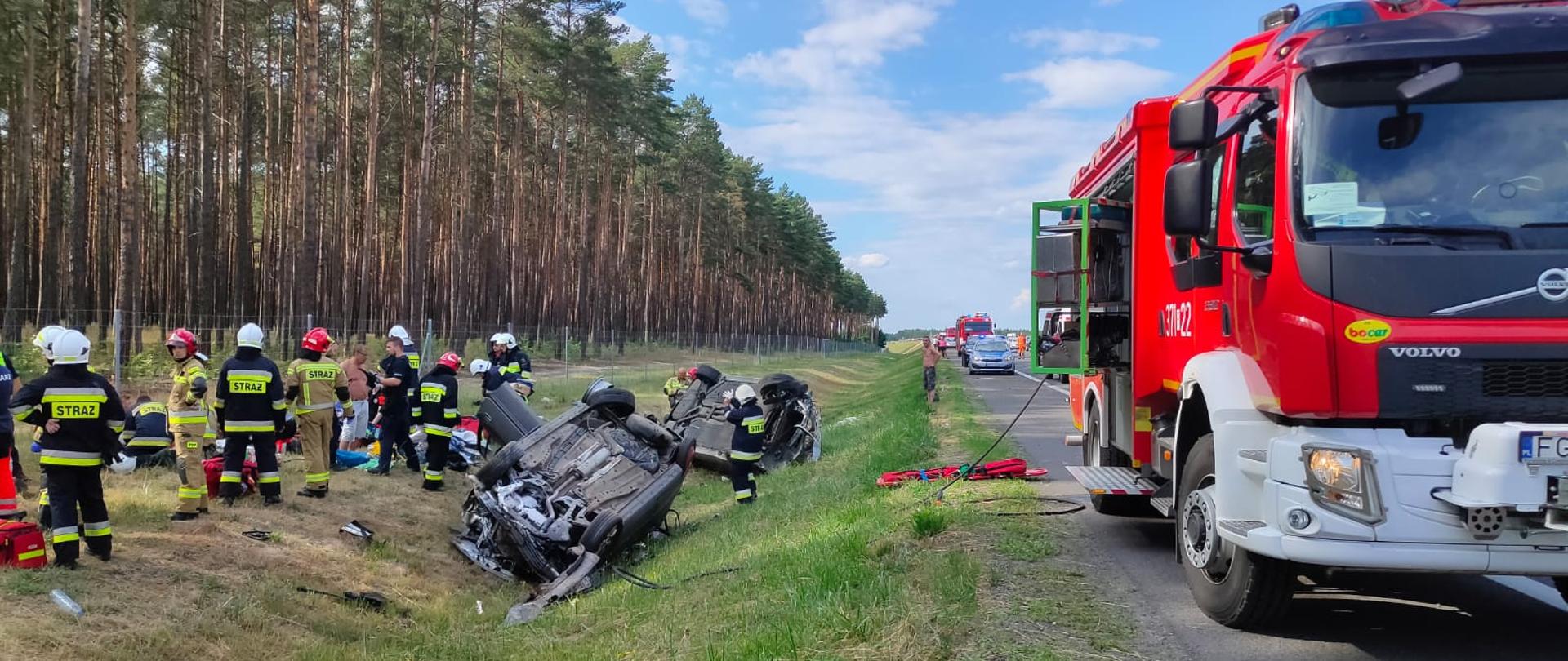 Zdjęcie przedstawia wypadek dwóch pojazdów osobowych na S-3, w którym uczestniczyło 9 poszkodowanych. Oba pojazdy leżą kołami do góry w rowie przy drodze. Strażacy zaopatrują poszkodowanych leżących w punkcie medycznym na nasypie przy drodze.