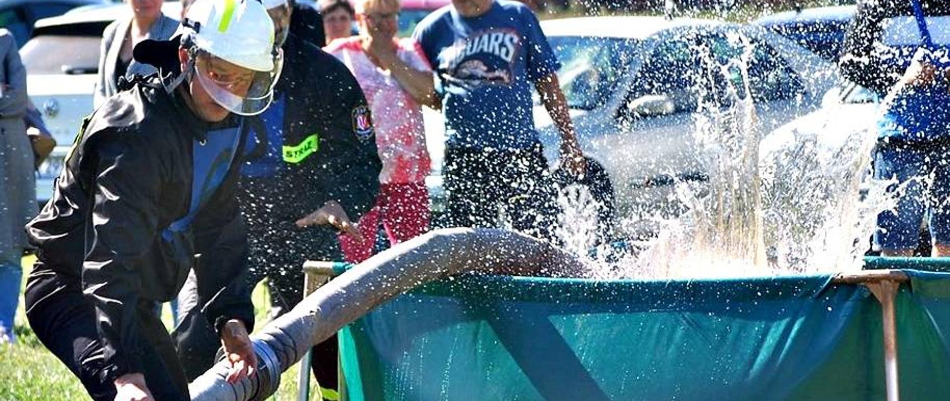 z lewej strony dwóch strażaków OSP w ubraniu koszarowym wrzucają linię ssawną do zbiornika brezentowego wypełnionego wodą.