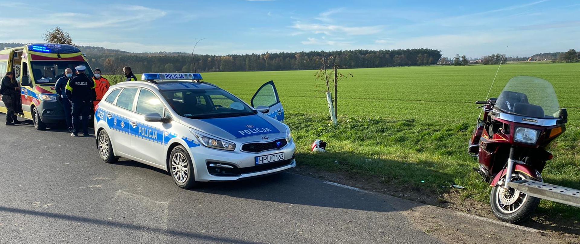 Po prawej stronie stoi motocykl biorący udział w zdarzeniu. Na głównym planie radiowóz policyjny na tle pola, a po lewej stronie ZRM zaopatrujący kierowcę jednośladu.