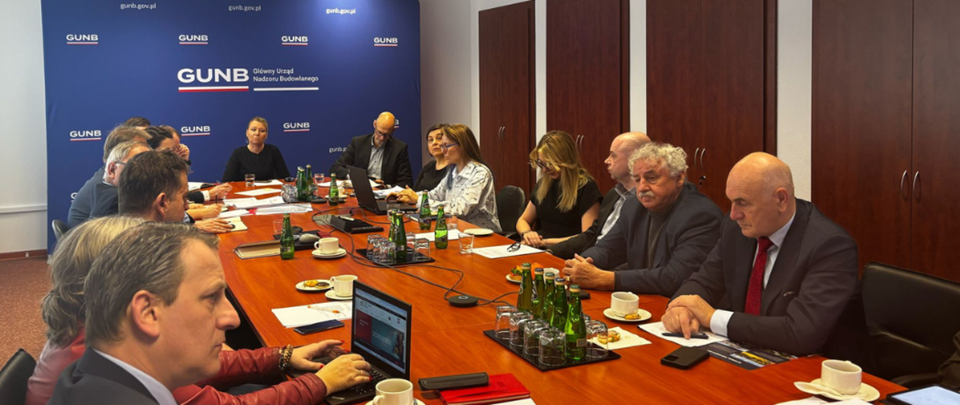 Uczestnicy posiedzenia Rady Doradczej Głównego Inspektora Nadzoru Budowlanego siedzący przy długim stole konferencyjnym. W tle ścianka z logo GUNB. Na stole leżą dokumenty, laptopy, butelki z wodą i filiżanki z kawą.
