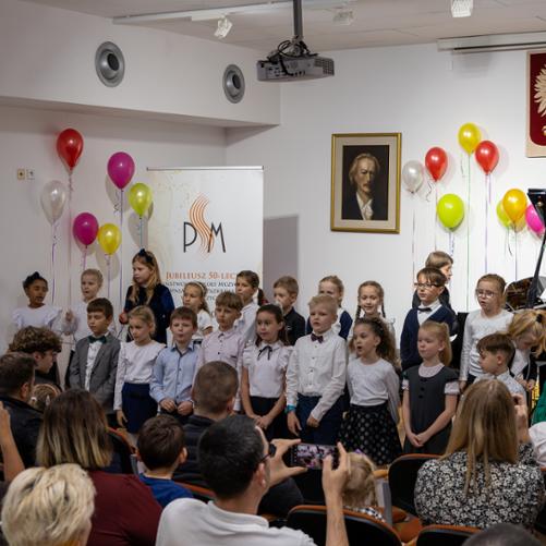 Pierwszoklasiści śpiewają wspólnie na scenie PSM w Głubczycach podczas uroczystości pasowania na ucznia. Przy fortepianie pani Małgorzata Meyse. W dolnej części zdjęcia widoczna publiczność.