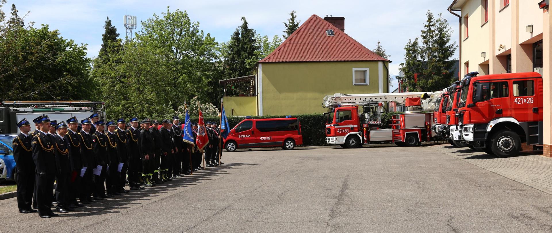 Pododdział stoi w dwuszeregu, za nim poczty towarzyszące. W tle samochody pożarnicze.