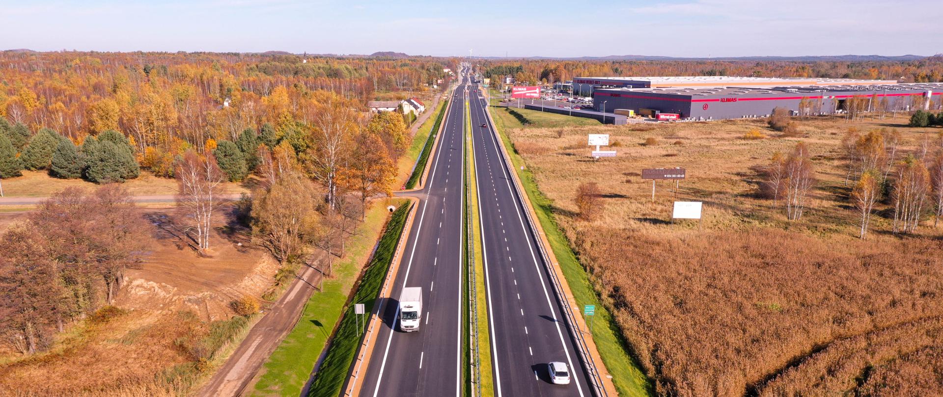 Dwujezdniowa droga krajowa po dwa pasy ruchu w obu kierunkach przebiegająca między lasami i budynkami magazynowymi. Zdjęcie wykonane późną jesienią. Drzewa pozbawione częściowo liści, miejscami przebijający złoty i czerwony kolor drzew.