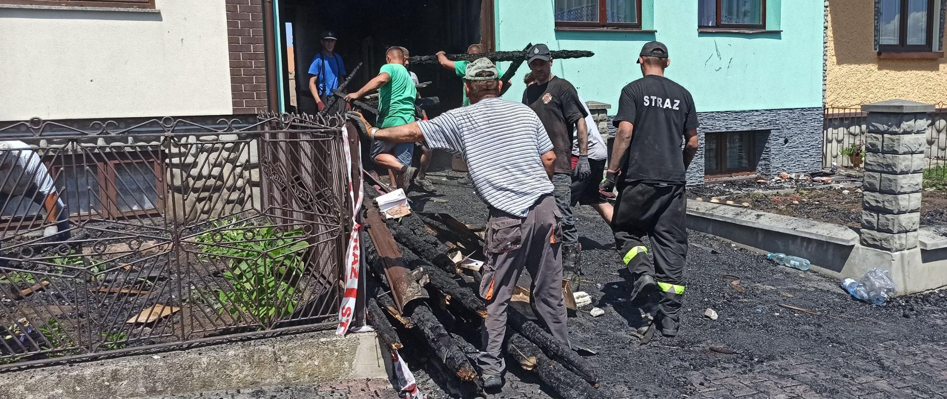Strażacy i mieszkańcy porządkują teren przed budynkiem, który uległ częściowemu spaleniu.
