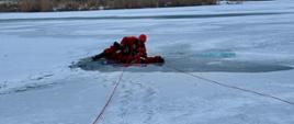 Ratownik leży na lodzie przy przeręblu, stabilizując osobę wyciągniętą z lodowatej wody podczas ćwiczeń ratownictwa lodowego na zamarzniętym zbiorniku.