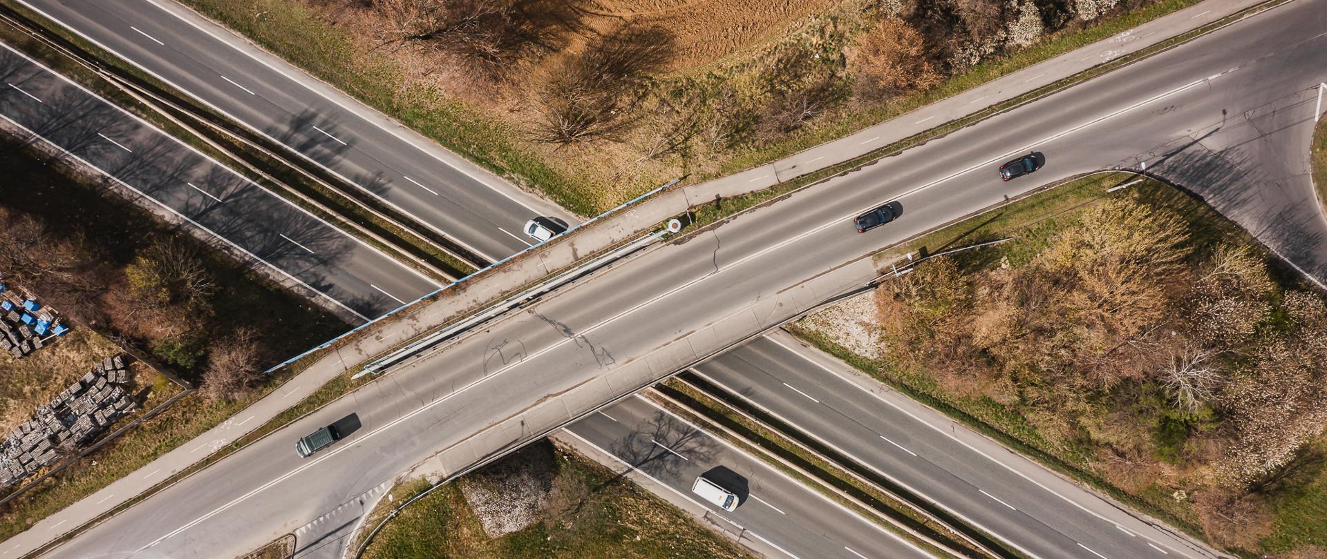Zdjęcie przedstawia wiadukt nad drogą krajową DK86 w Będzinie. Wiadukt przecina ruchliwą trasę, umożliwiając przejazd nad nią. Na zdjęciu widoczne są pojazdy poruszające się po różnych pasach ruchu, a także obszary zieleni z drzewami po jednej stronie oraz materiały budowlane po drugiej. Ujęcie z góry pozwala dostrzec organizację ruchu oraz infrastrukturę drogową w tym miejscu.