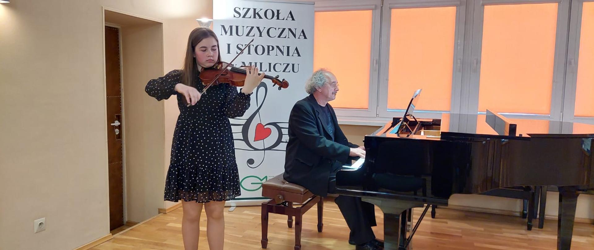 Dziewczynka stoi, trzyma skrzypce, nauczyciel siedzi przy fortepianie