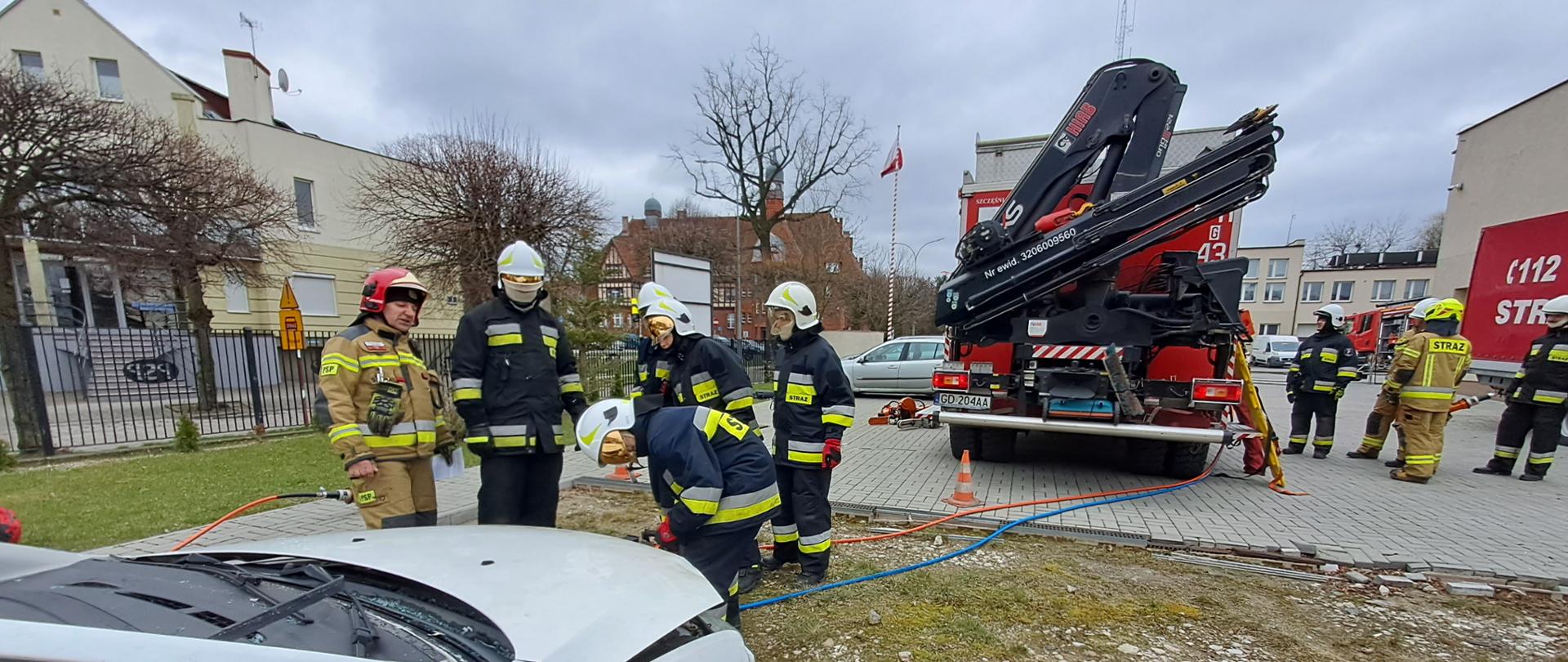 Stanowisko egzaminacyjne z ratownictwa technicznego. Strażak w ubraniu specjalnym z zamkniętą przyłbicą pracuje rozdzielaczem hydraulicznym na wraku pojazdu, obok instruktor i inni kandydaci na strażaków.