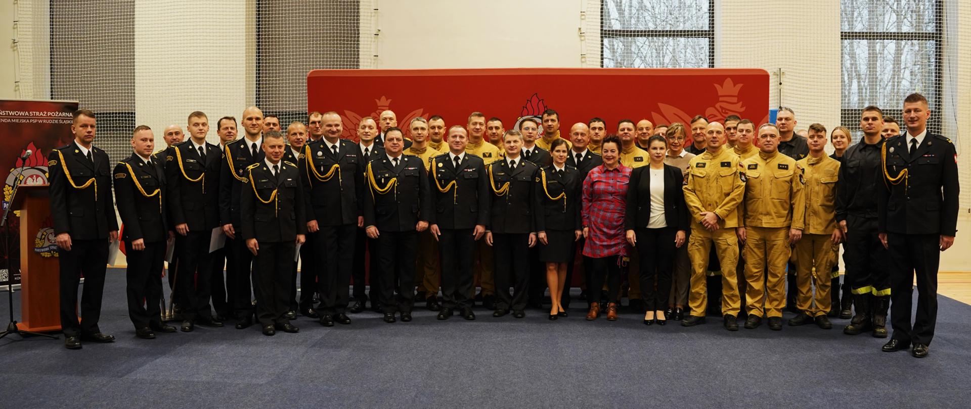 Duża grupa strażaków w umundurowaniu galowym i służbowym oraz pracowników cywilnych stoi razem w hali na tle czerwonej ścianki z emblematem PSP, pozując do wspólnego zdjęcia.