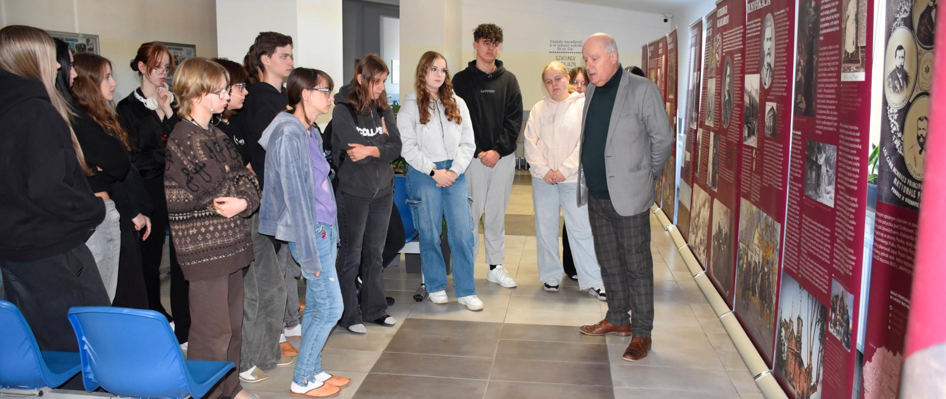 Przed wystawą stoi nauczyciel mówiąc do zgromadzonej młodzieży