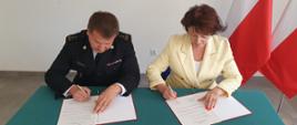 Na zdjęciu Komendant Powiatowy PSP w Gorlicach w umundurowaniu wyjściowym granatowym podpisuje porozumienie obok siedzi Pani Dyrektor Powiatowej Stacji Sanitarno-Epidemiologicznej w Gorlicach która również podpisuje porozumienie. Pani Dyrektor ubrana w kremowy żakiet i białą spódnicę. W.w osoby na tle ściany koloru białego po prawej stronie stoją biało czerwone flagi.