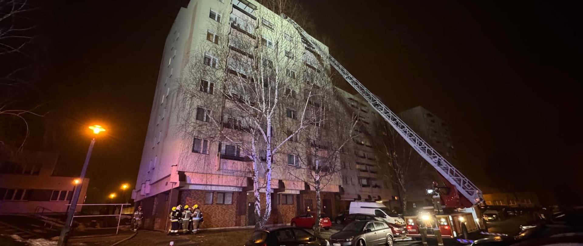 Nocna akcja straży pożarnej przy wysokim bloku mieszkalnym — wóz strażacki z wysuniętą drabiną sięga górnych pięter budynku, na dole stoją strażacy w ubraniach ochronnych, obok zaparkowane samochody i oświetlona latarniami mokra ulica.