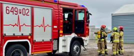 Przy dużym czerwonym wozie strażackim z numerem 649[P]02 stoi kilku strażaków w pełnym umundurowaniu ochronnym. Pojazd ma otwarte drzwi kabiny i zamknięte skrytki sprzętowe z boku. Na burcie widać ozdobny wzór w kształcie linii EKG. Strażacy omawiają działania, stojąc w grupie obok pojazdu, na tle szarego budynku.