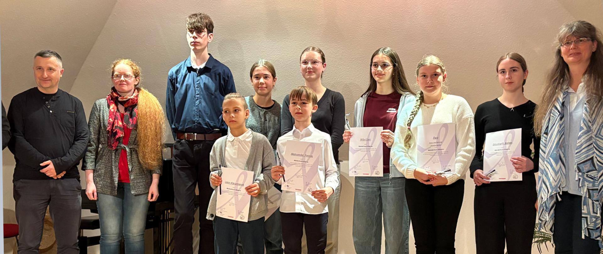 Grupa kilkunastu osób, uczniowie i nauczyciele stoją na tle ściany, dzieci w rękach trzymają dyplomy.