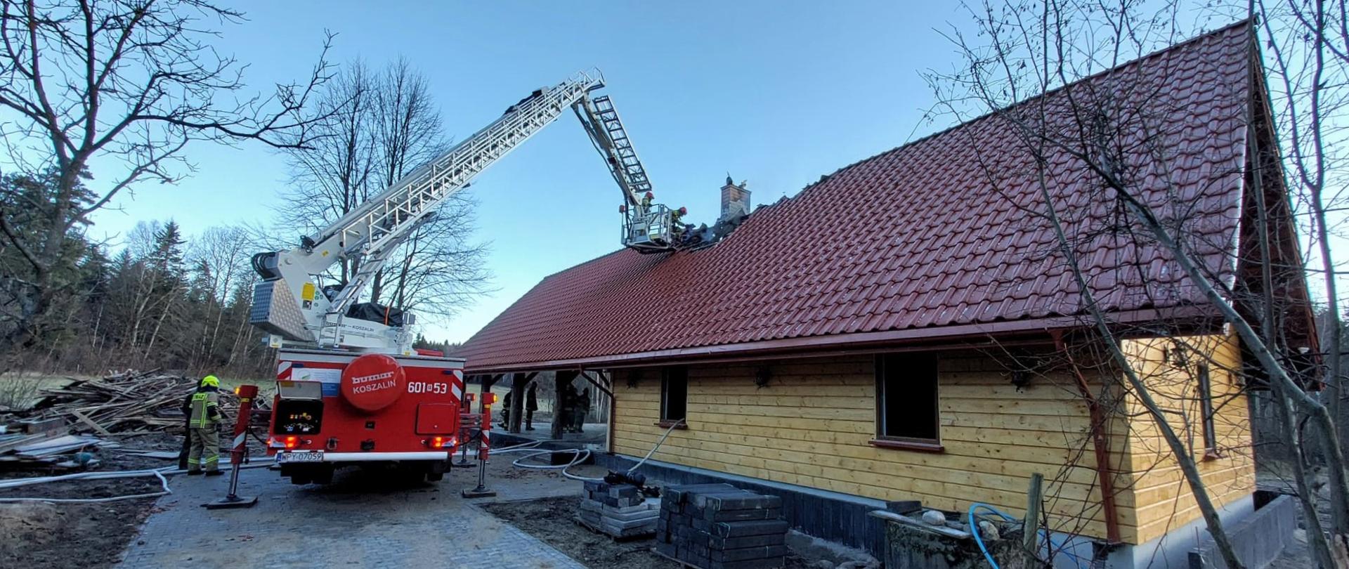 Zdjęcie przedstawia akcję gaśniczą na dachu budynku mieszkalnego w ciągu dnia, prawdopodobnie po pożarze komina lub dachu. Na pierwszym planie po lewej stronie znajduje się czerwony wóz strażacki z numerem "601 53" na boku. Od wozu odchodzi długa, biała drabina hydrauliczna z koszem na końcu, który jest uniesiony i znajduje się nad dachem drewnianego domu. W koszu drabiny widać strażaków, którzy operują na wysokości. Dom ma drewniane ściany i spadzisty dach pokryty czerwoną dachówką. Przed domem, na ziemi, widoczne są fragmenty sprzętu gaśniczego, w tym węże strażackie. W pobliżu wozu strażackiego stoją dwaj strażacy w żółtych mundurach, obserwując sytuację. Tło zdjęcia stanowią nagie drzewa liściaste i iglaste, co sugeruje wczesną wiosnę lub późną jesień. Niebo jest jasne i bezchmurne. Po lewej stronie widać fragment podjazdu z kostki brukowej i stosy drewna lub gruzu. Całość obrazu wskazuje na działania straży pożarnej w celu zabezpieczenia lub usunięcia skutków zdarzenia.