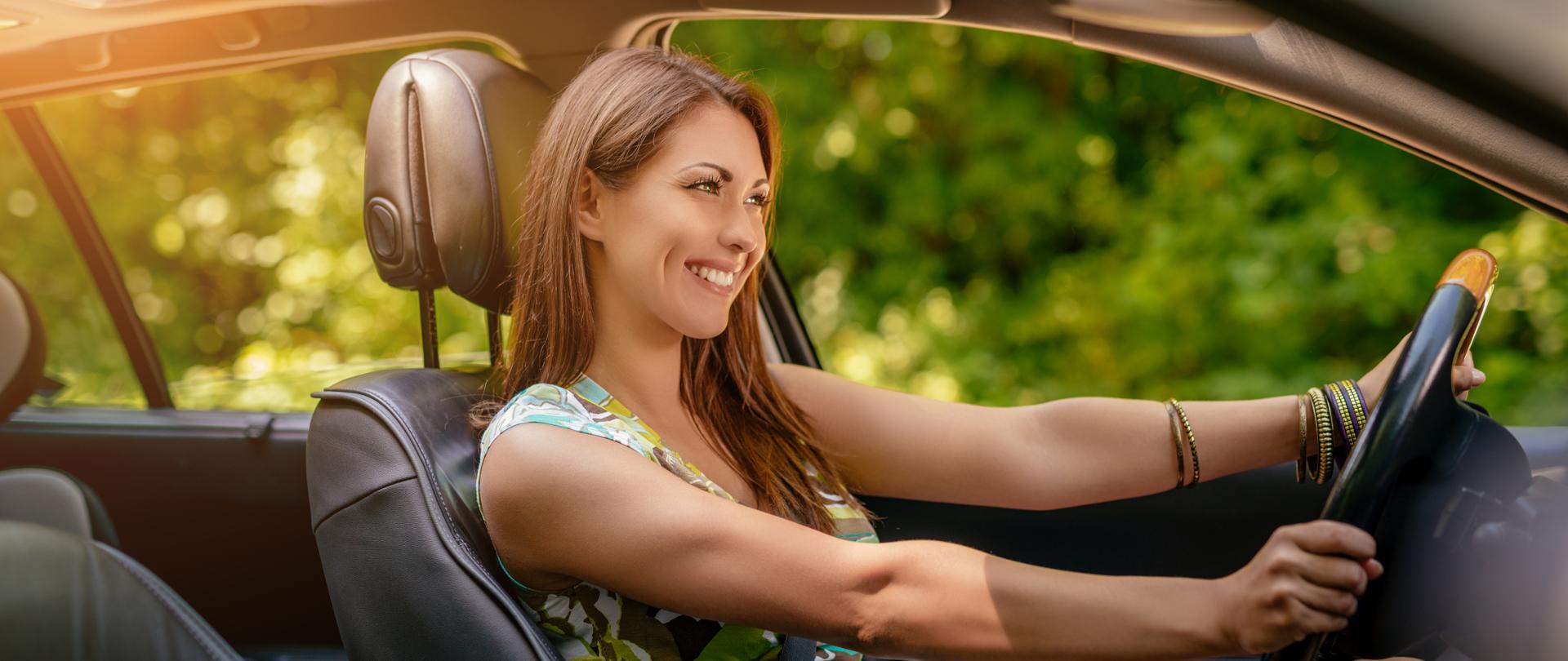 Na zdjęciu młoda, uśmiechnięta kobieta za kierownicą samochodu. 