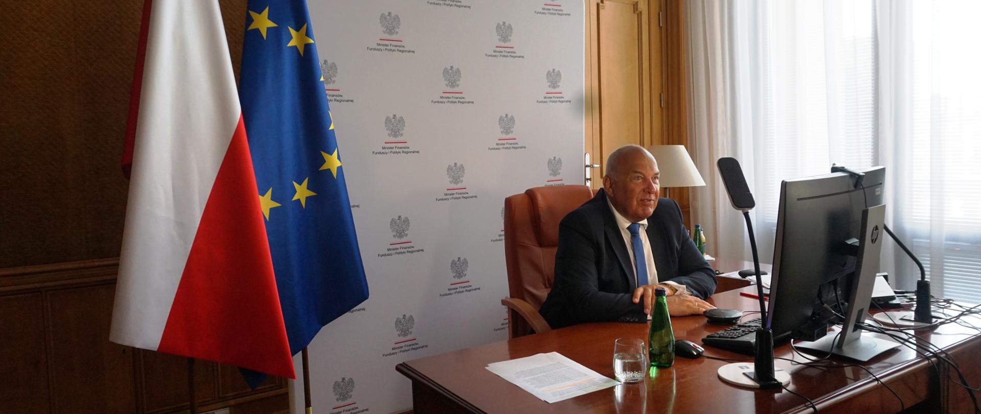 W gabinecie przy biurku siedzi Tadeusz Kościński. Przed nim ekran monitora. Za nim flagi PL i UE oraz ścianka Ministerstwa Finansów.