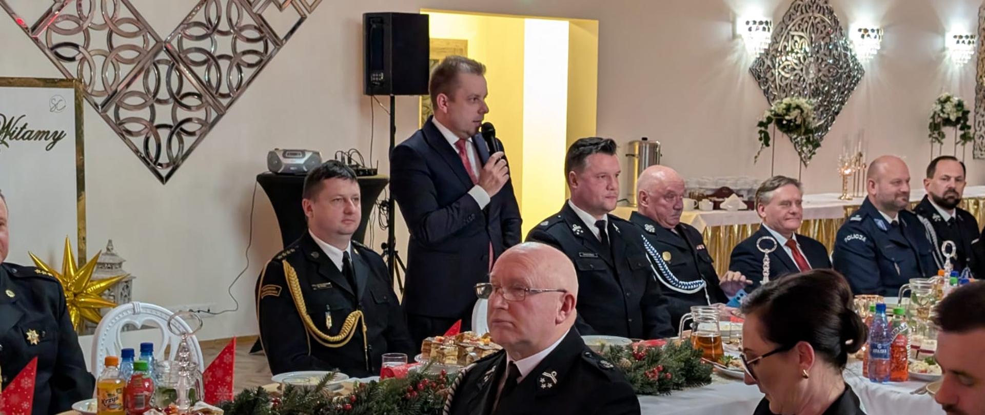 Uroczyste spotkanie wigilijne w sali bankietowej. Przy długim, świątecznie udekorowanym stole siedzą przedstawiciele służb mundurowych w galowych i służbowych mundurach oraz osoby cywilne. Mężczyzna w garniturze stoi i przemawia do zebranych, trzymając mikrofon. Na stole widoczne są potrawy wigilijne, ciasta, napoje, świece oraz dekoracje z gałązek świerkowych i czerwonych serwetek. W tle jasna sala z eleganckimi zdobieniami ściennymi i oświetleniem.