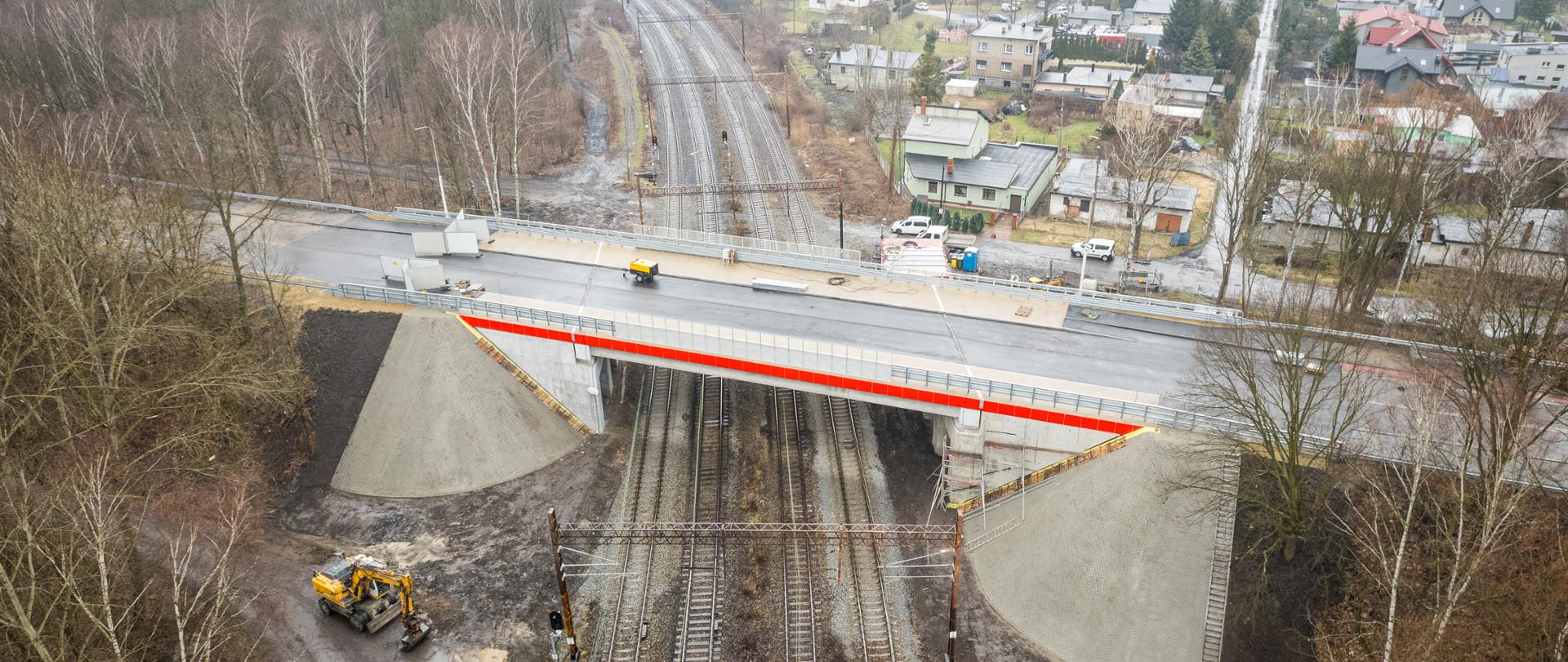 Wiadukt nad torami kolejowymi, prawej stronie znajdują się budynki mieszkalne, a po lewej stronie drzewa. 
