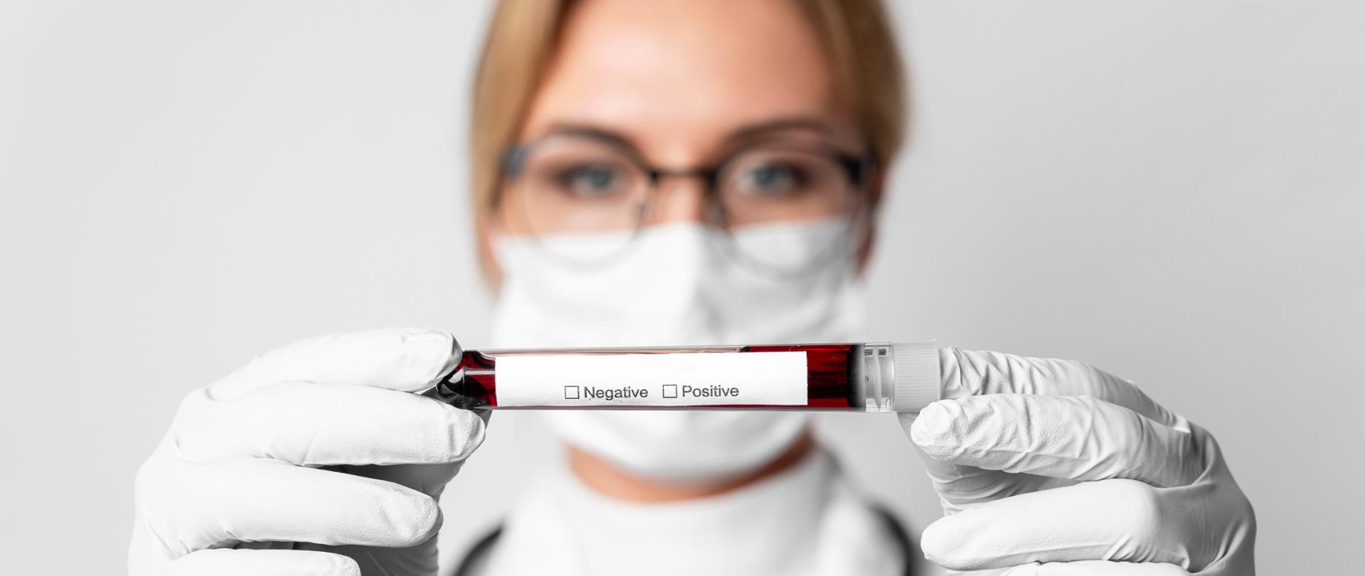 kobieta w maseczce i odzieży ochronnej trzymająca w ręku fiolkę z pobraną krwią na test na HIV