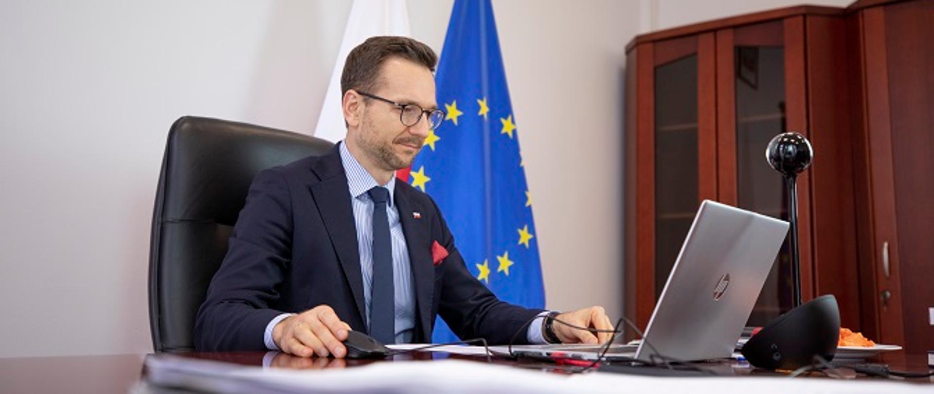 Wiceminister Waldemar Buda siedzi przy biurku przed laptopem. Za plecami stoją flagi Polski i Unii Europejskiej.