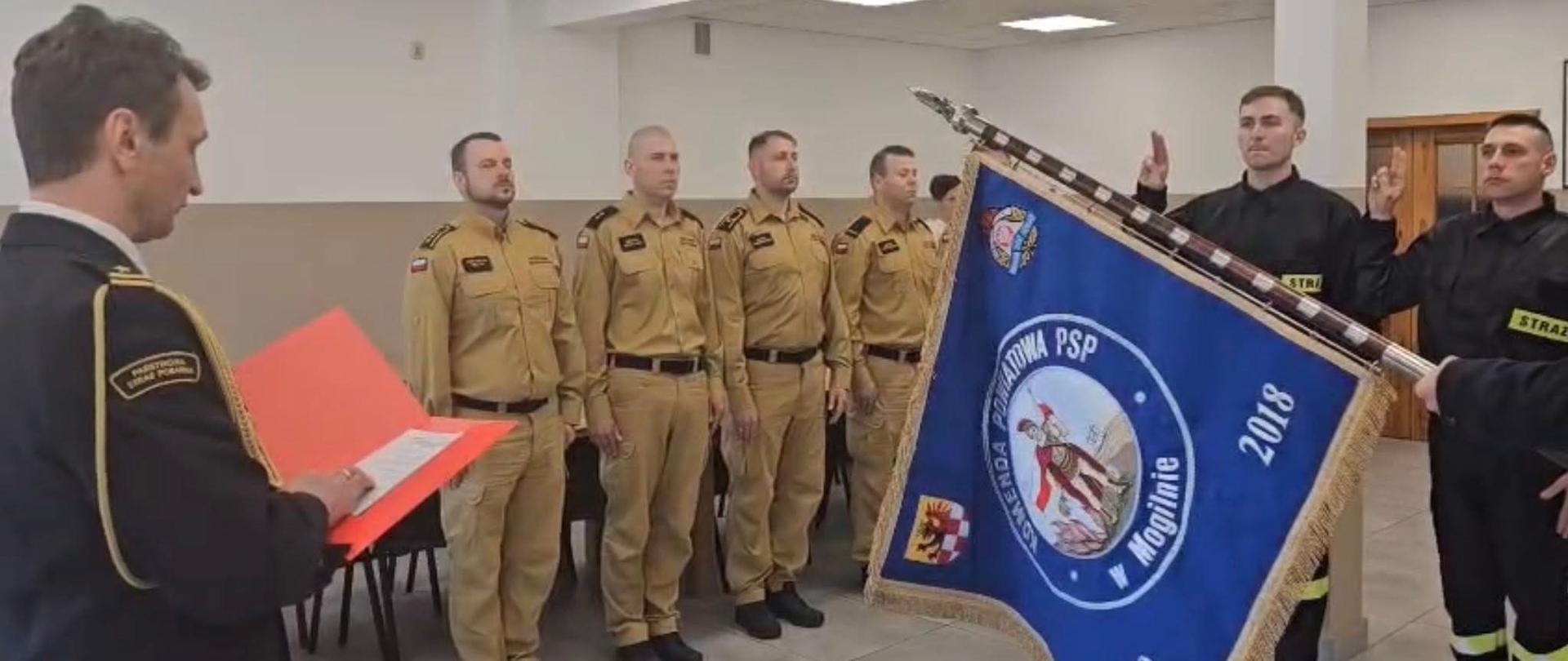 Nowo przyjęci strażacy składają ślubowanie. Poczet sztandarowy stoi z opuszczonym sztandarem. Po lewej stronie stoją funkcjonariusze. Komendant trzyma w ręku kartkę z tekstem roty ślubowania.