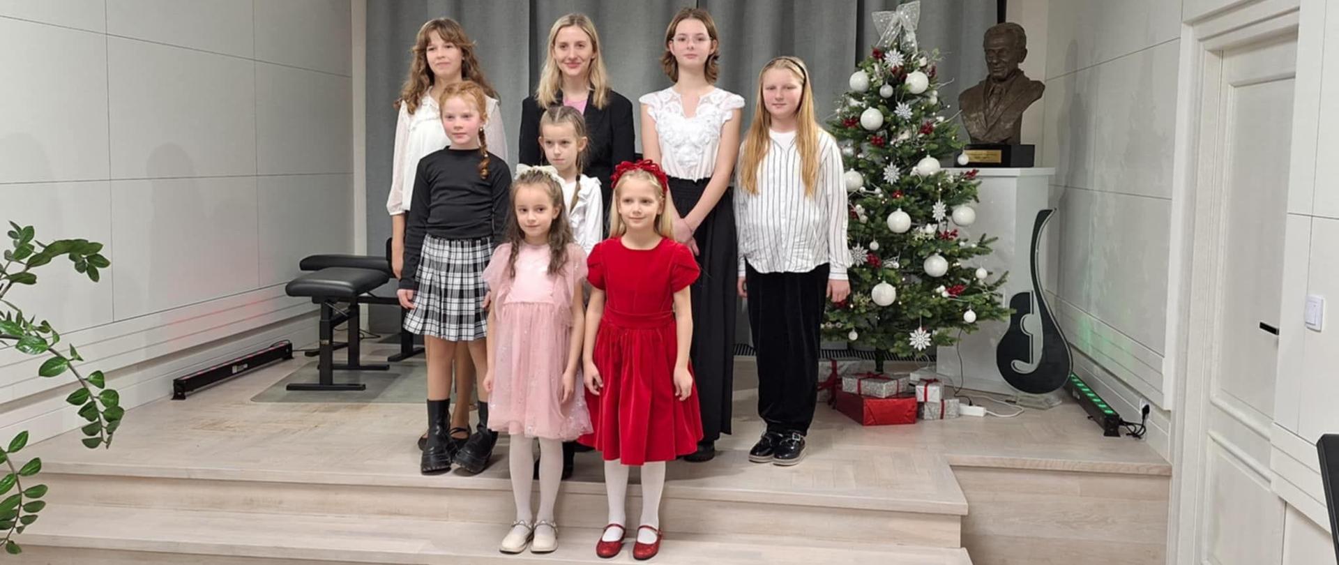 Na zdjęciu dziewczynki z nauczycielem stoją w dwóch rzędach na niewielkim podium. Są elegancko ubrano a obok stoi przystrojona choinka z prezentami. Pomieszczenie jest nowoczesne, z jasnym wykończeniem wnętrza. Po lewej stronie znajduje się roślina doniczkowa, po prawej – popiersie patrona oraz statuetka szklana przypominająca kształtem gitarę albo klucz wiolinowy.