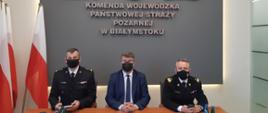 Po lewo flagi RP, na wprost siedzą od lewo komendant wojewódzki PSP w Białymstoku, sekretarz stanu w MSWiA Maciej Wąsik oraz komendant główny PSP, w tle napis na szarym tle Komenda Wojewódzka PSP w Białymstoku