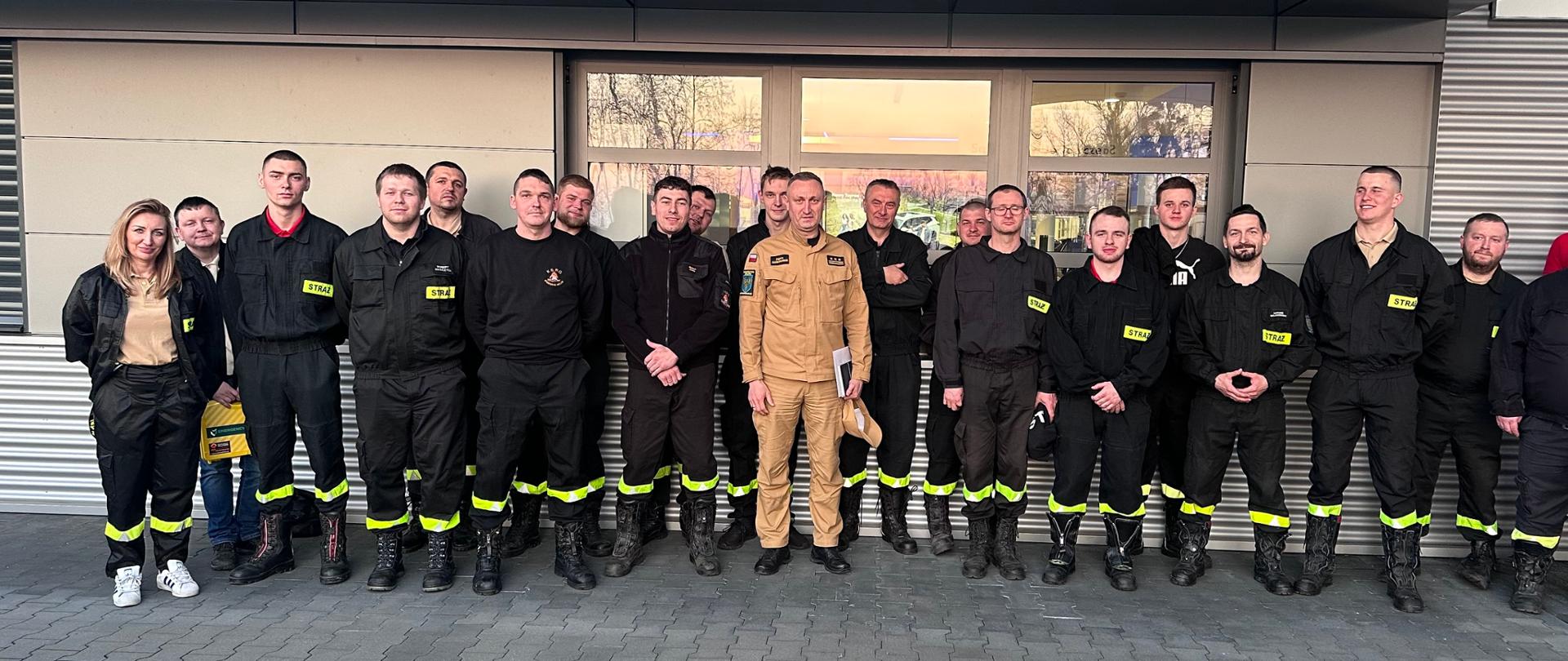 Na zdjęciu znajdują się strażacy w umundurowaniu koszarowym ustawieni do zdjęcia przed budynkiem