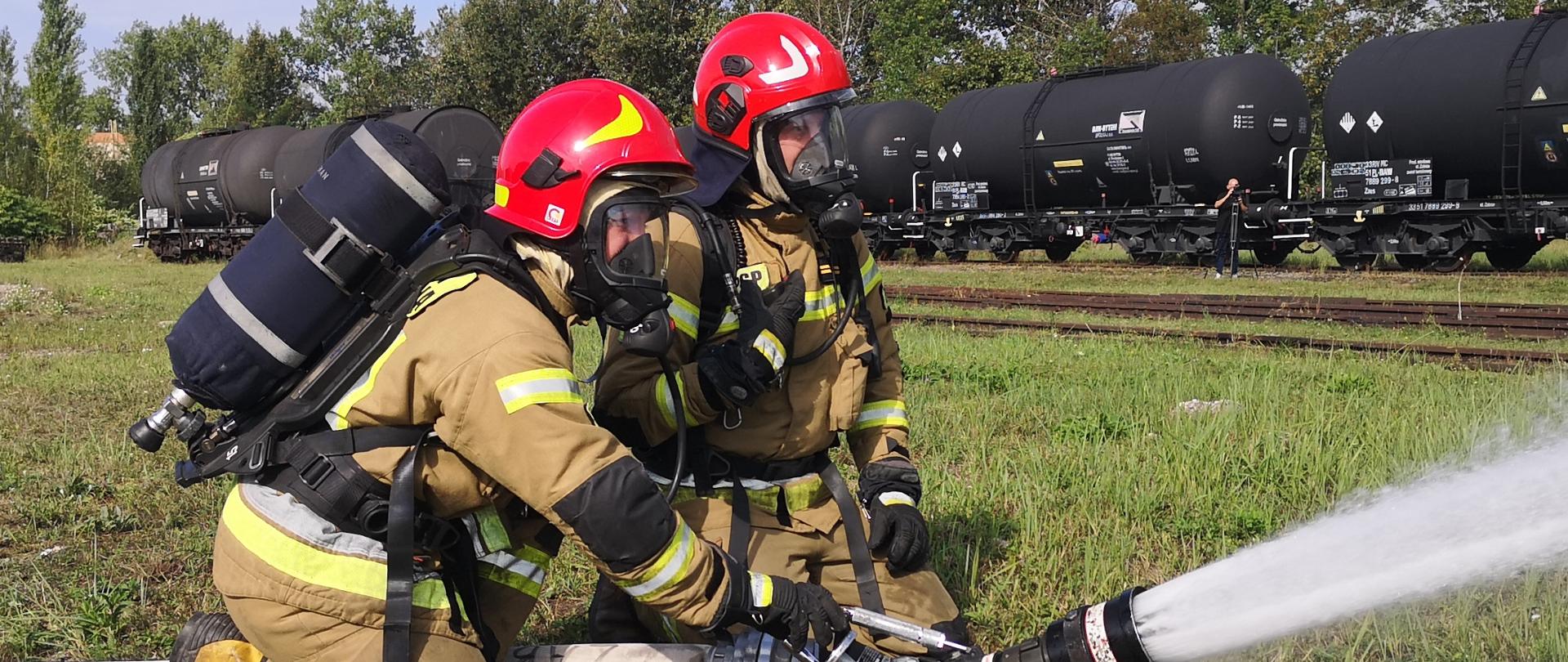 Na zdjęciu dwaj strażacy wyposażeni w aparaty ochrony dróg oddechowych operują działkiem wodnym. W tle widoczne cysterny kolejowe, drzewa, niebo częściowo zachmurzone. 