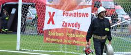 Zdjęcie przedstawia strażaka niosącego rozdzielacz i węża, na tle baner X powiatowych zawodów. sportowo-pożarniczych OSP