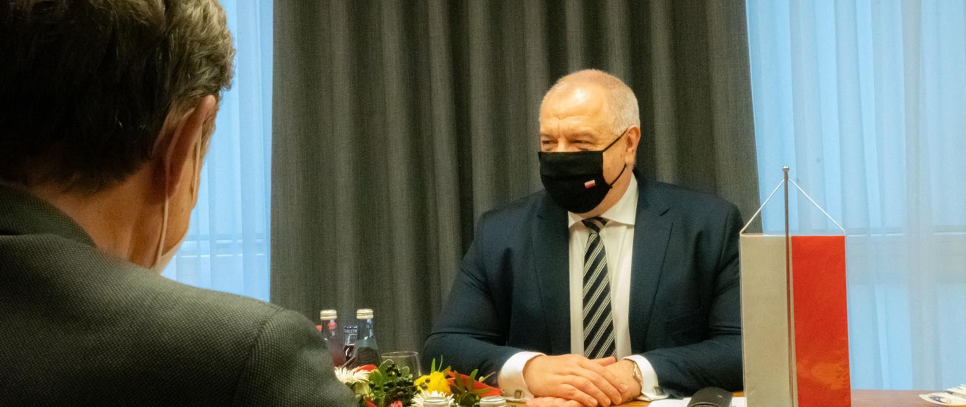 Ujęcie zza ramienia ambasadora. Wicepremier Jacek Sasin siedzi w maseczce naprzeciw swojego rozmówcy. Trzyma dłonie złożone na blacie. Po prawej stronie stoi mała polska flaga.