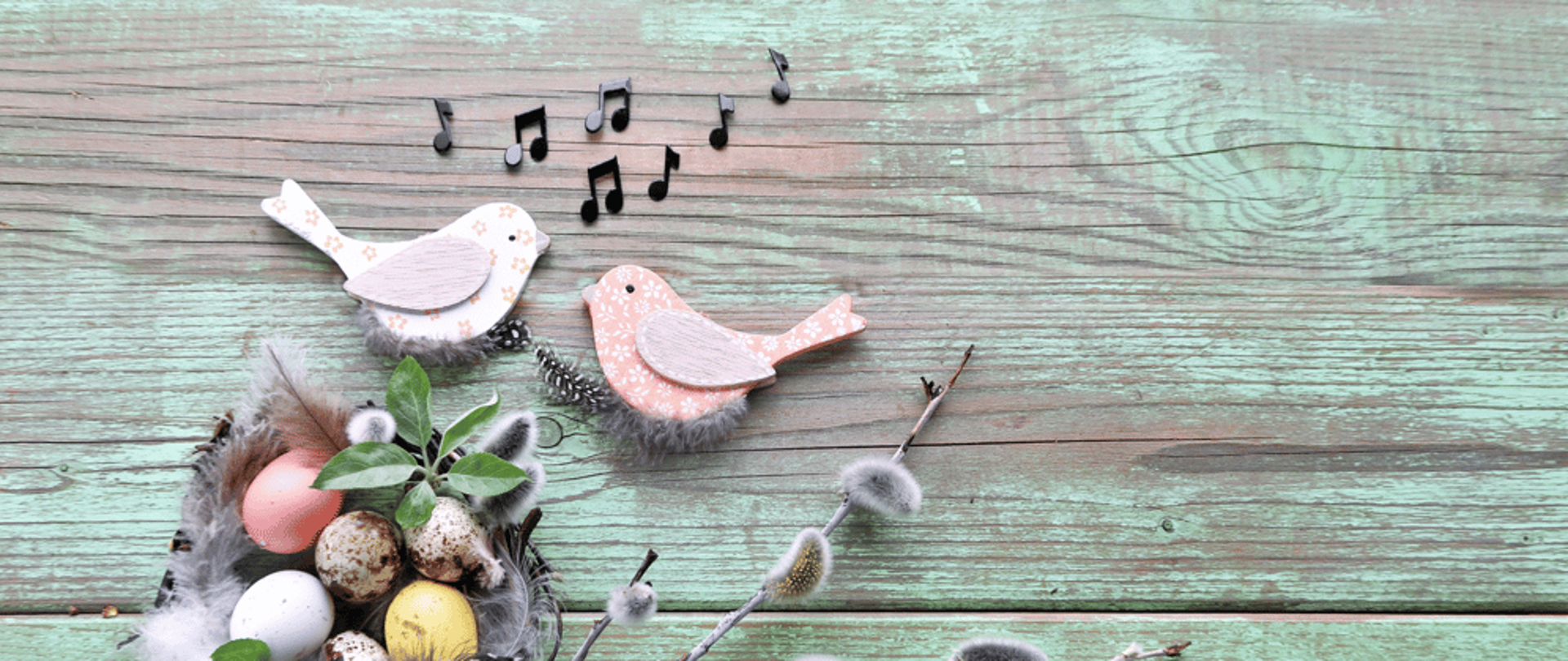 zdjęcie przedstawiające koszyczek z pisankami na zielonkawej desce. U góry dwa drewniane ptaszki oraz czarne nutki