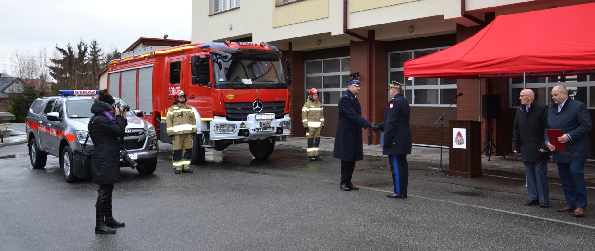 KomendantPowiatowy st. bryg. Witold Wiercioch odbiera od nadbryg. Andrzeja Babca, akt przekazania, w tle dwa samochody.