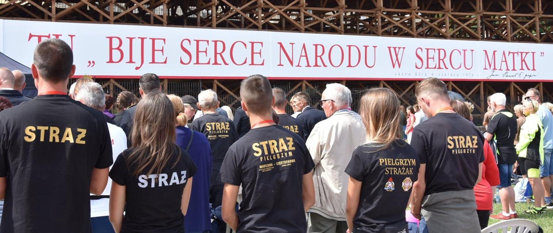 Osoby ze straży pożarnej stoją tyłem przed napisem Tu bije serce narodu w sercu Matki 