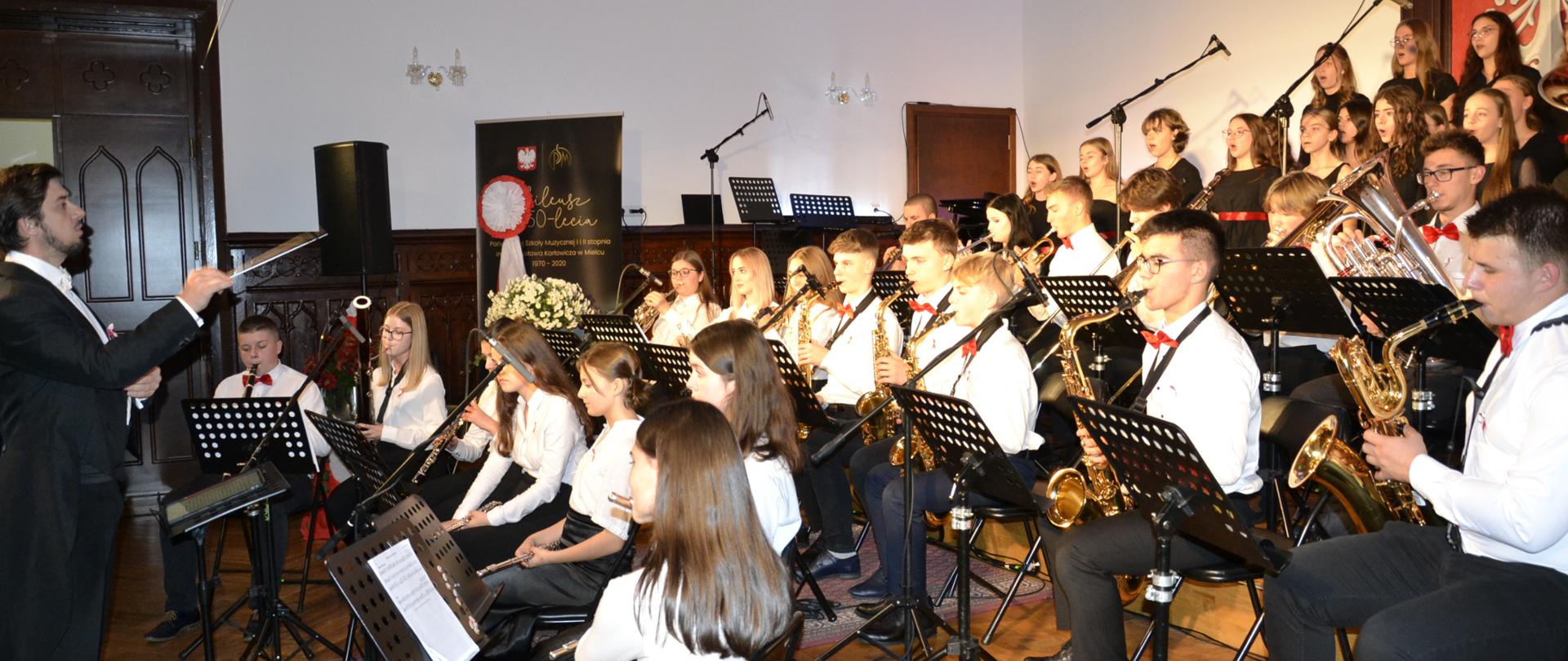 zdjęcie przedstawia orkiestrę dętą PSM II stopnia w Mielcu w ujęciu z prawej strony, z dyrygentem Piotrem Rysiewiczem po lewej stronie zdjęcia, muzycy mają na sobie białe bluzki z biało-czerwonymi kotylionami celem upamietnienia rocznicy odzyskania przez Polskę niepodległości, w tle widać również chór