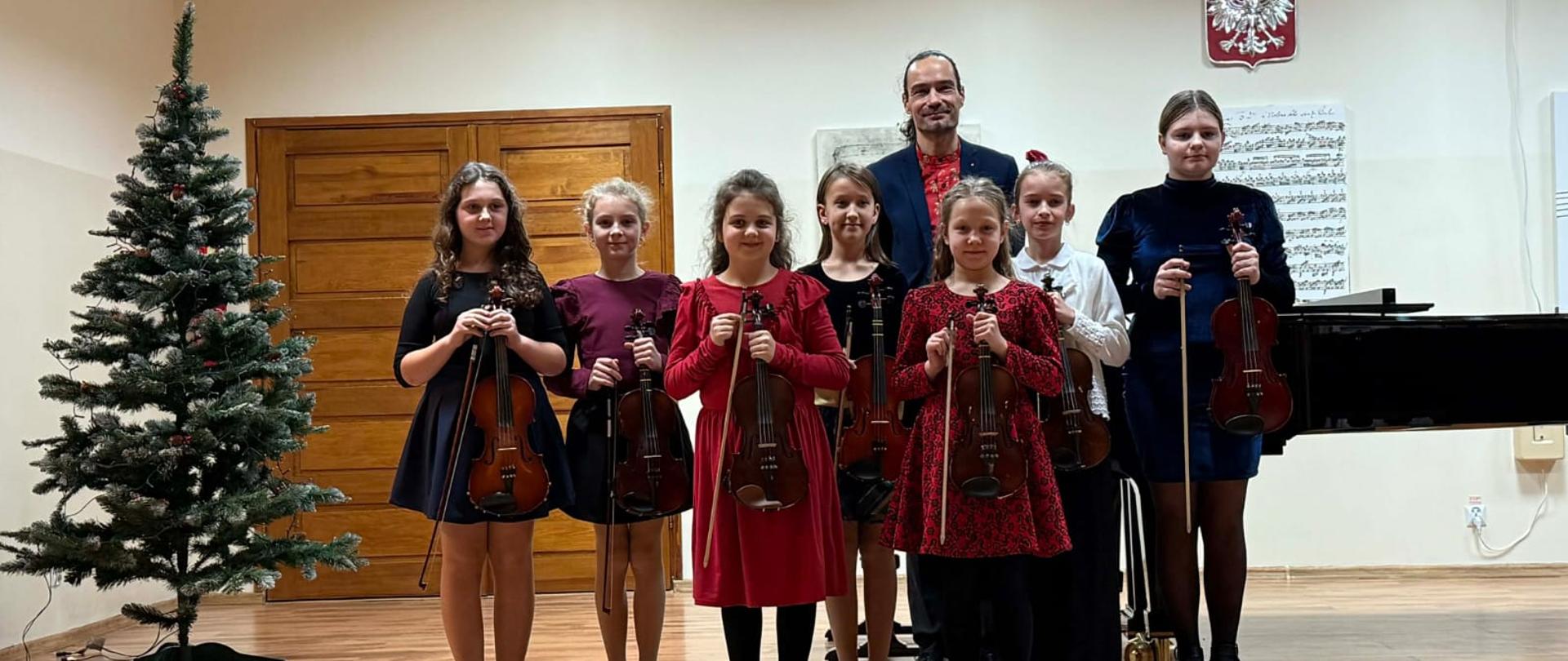 Zdjęcie przedstawia scenę a na niej grupa dzieci stojąca ze skrzypcami w rękach. Wśród nich nauczyciel. Dzieci i nauczyciel ubrani w czerwone i granatowe stroje. Po lewej stronie choinka, z tył widać czarny fortepian i godło Polski. Dzieci wraz z nauczycielem uśmiechają się