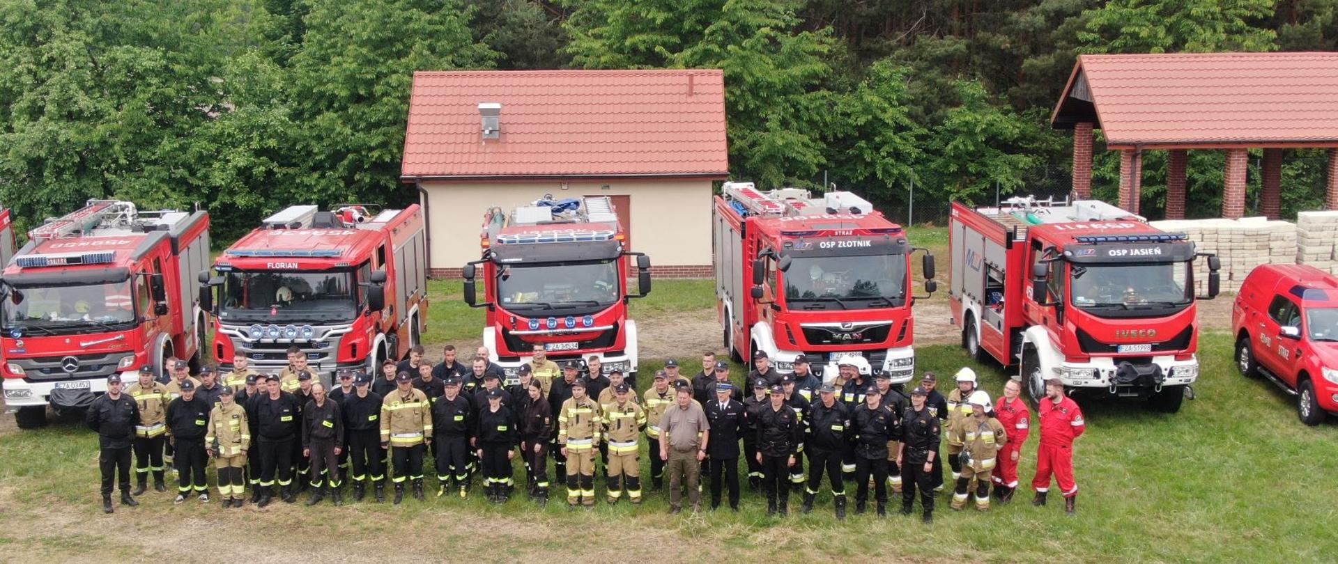 Strażacy w mundurach ustawieni przed pojazdami ratowniczo-gaśniczymi.