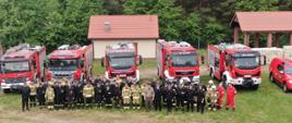 Strażacy w mundurach ustawieni przed pojazdami ratowniczo-gaśniczymi.